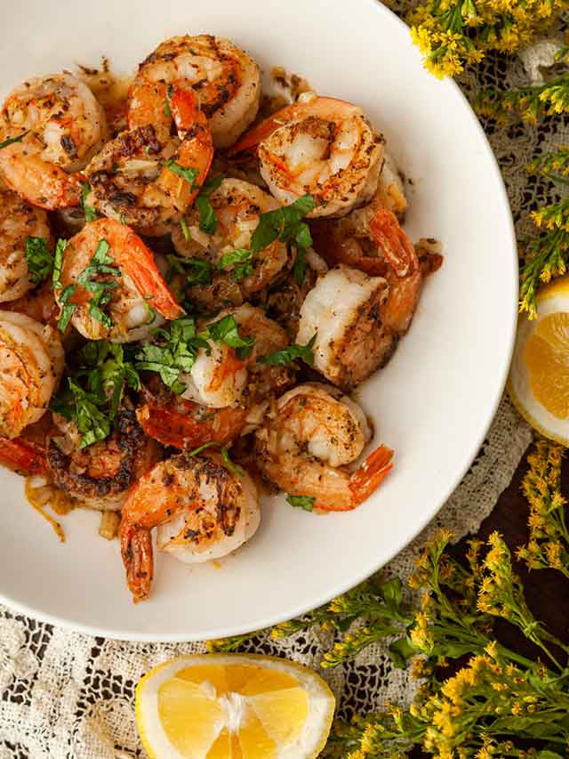 A bowl of lemon butter shrimp beside flowers and a lemon.