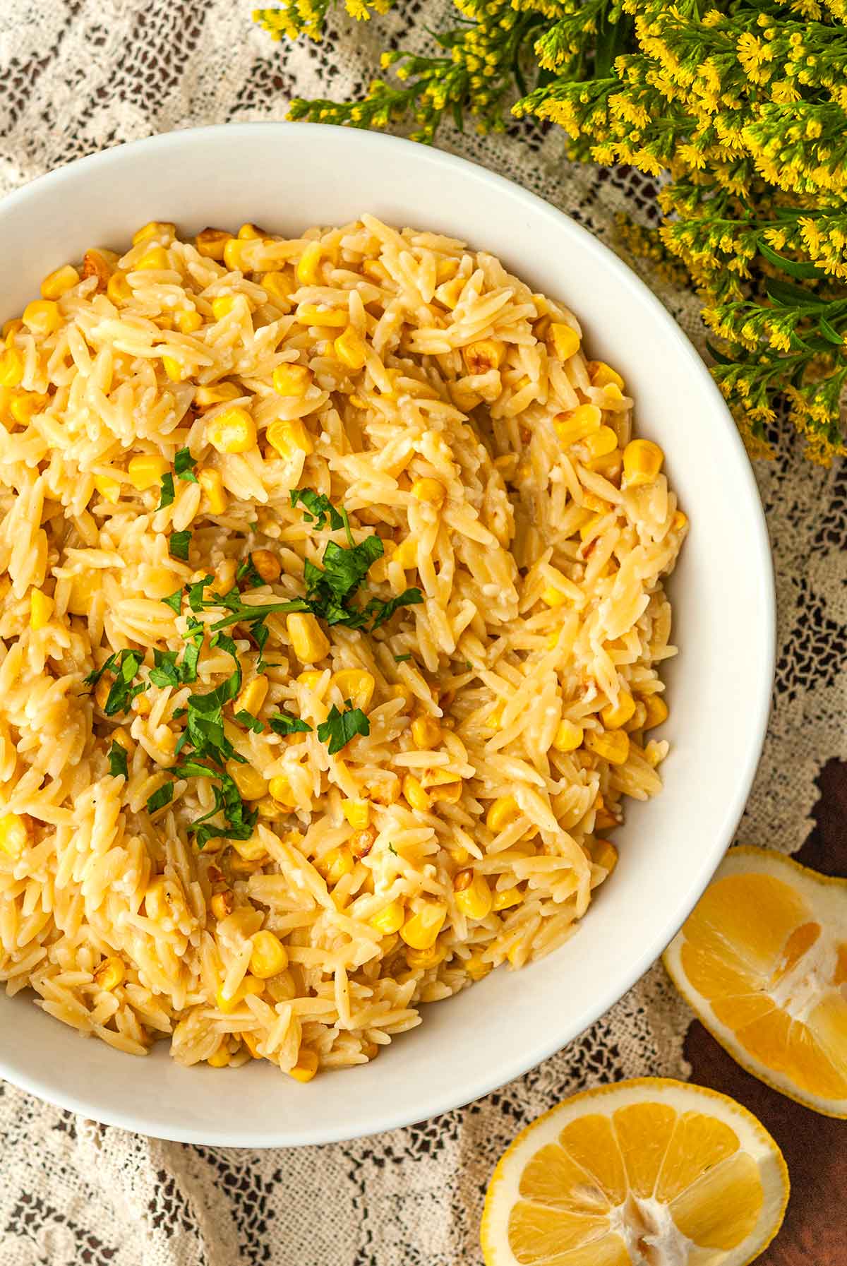A bowl of lemon orzo on a lace table cloth surrounded by lemon slices and flowers.