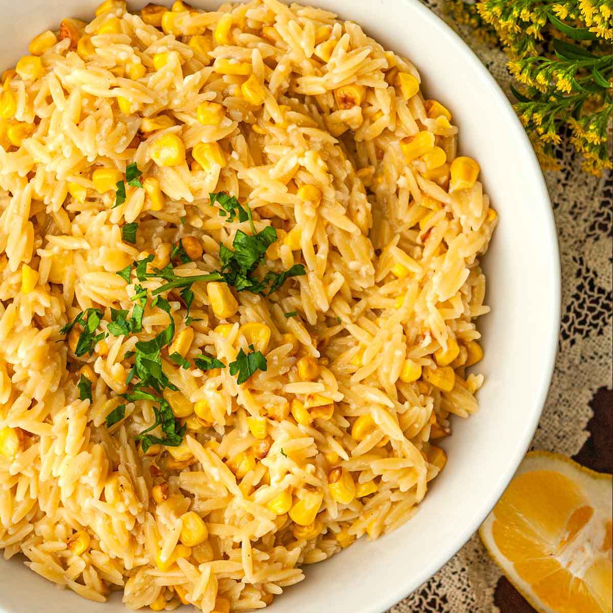 A bowl of lemon orzo on a lace table cloth surrounded by lemon slices and flowers.