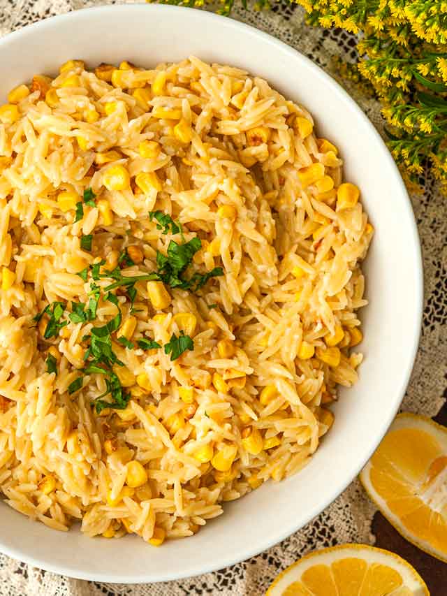 A bowl of lemon orzo on a lace table cloth surrounded by lemon slices and flowers.