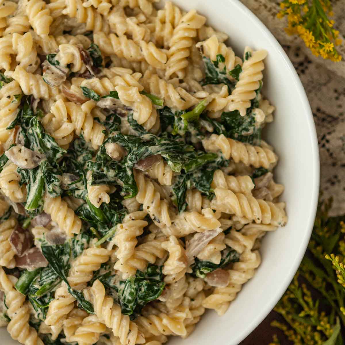 A bowl of pasta with spinach on a lace table cloth beside flowers