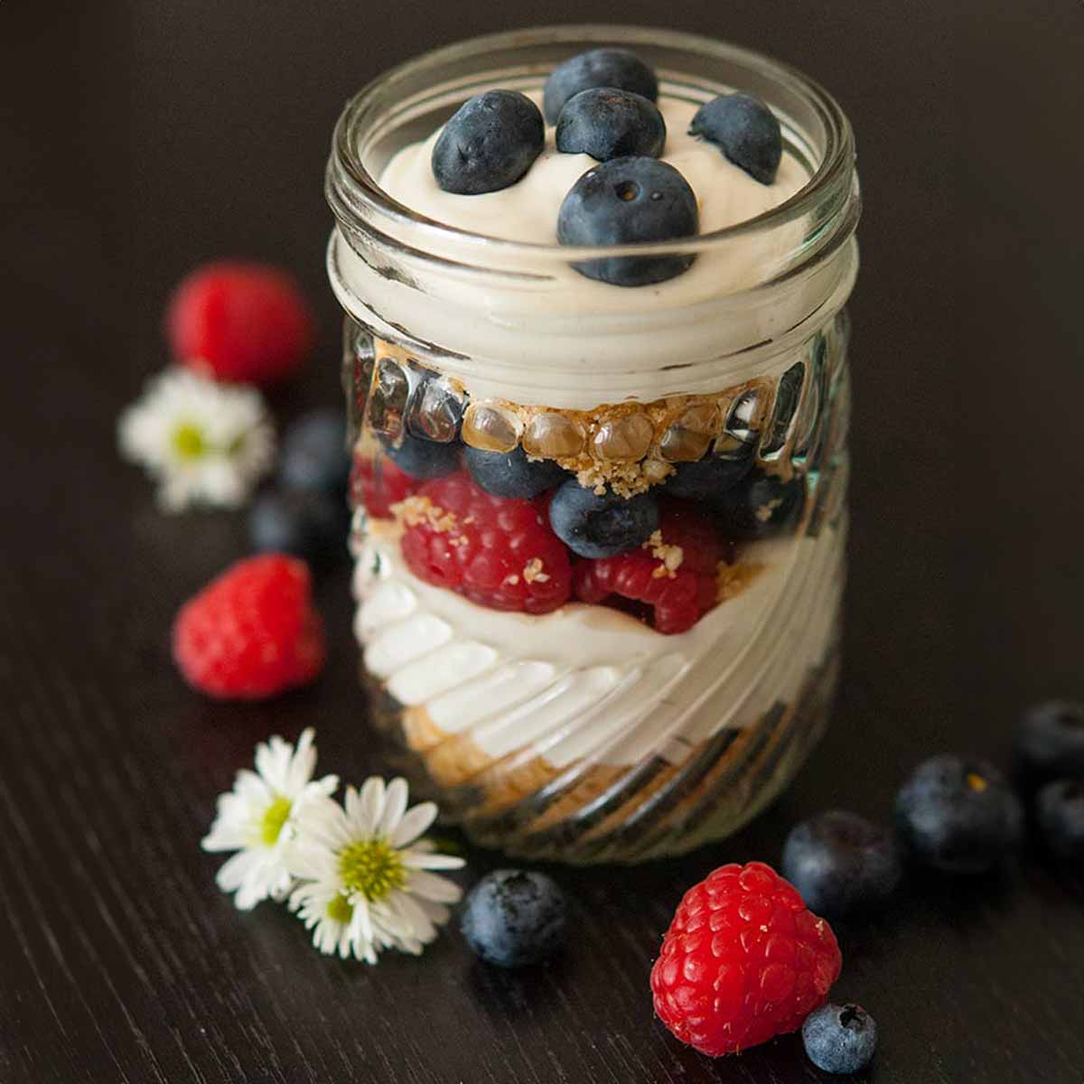 A mason jar with cheesecake mouse, cookie crumbles and berries on a black table, sprinkled with cookie crumbs and berries.