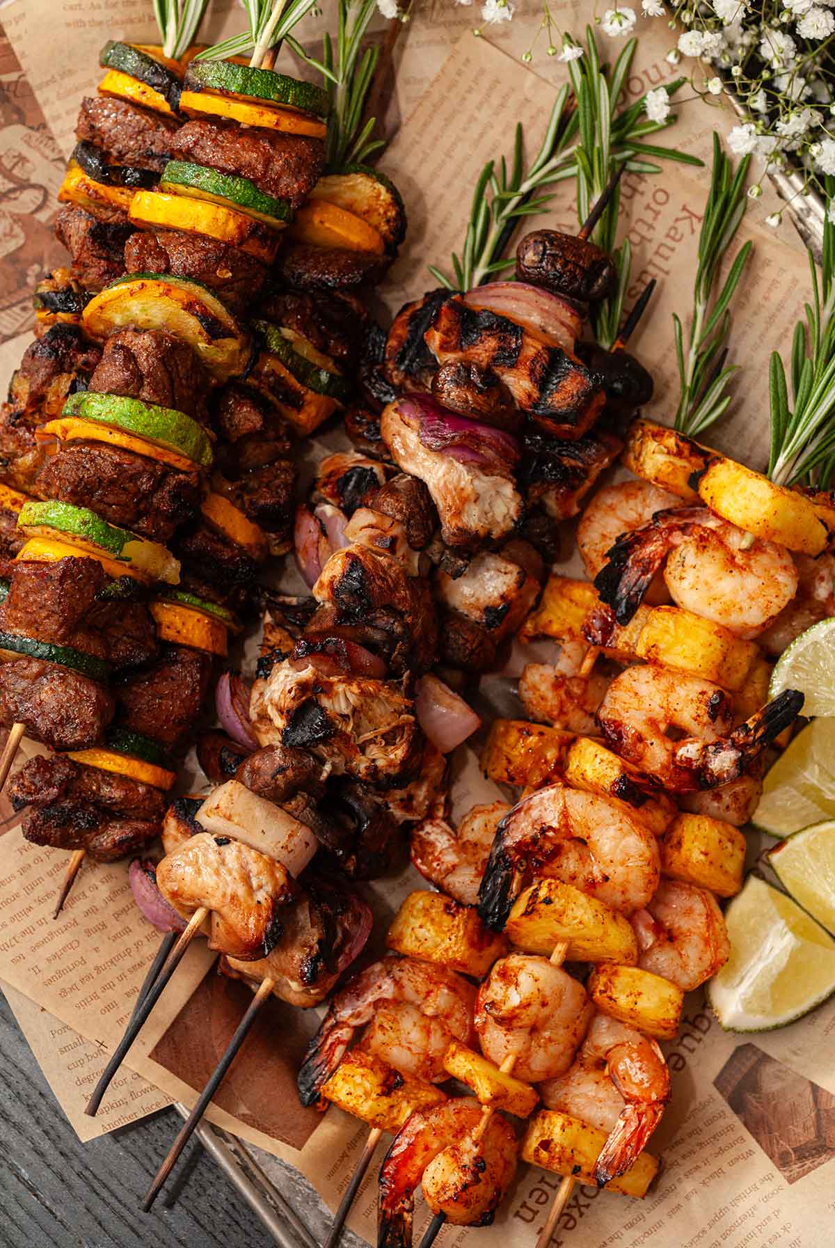 3 different kinds of meat, vegetable and fruit skewers on newsprint paper, with rosemary in the tops.