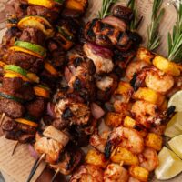 3 different kinds of meat, vegetable and fruit skewers on newsprint paper, with rosemary in the tops.