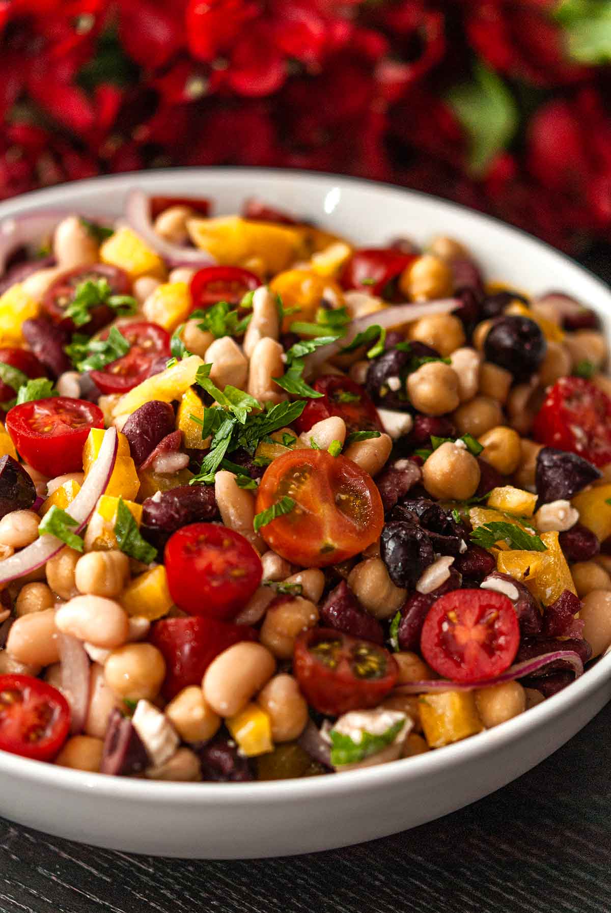 Tomatoes, beans, onion and feta in a bowl in front of flowers on a table.