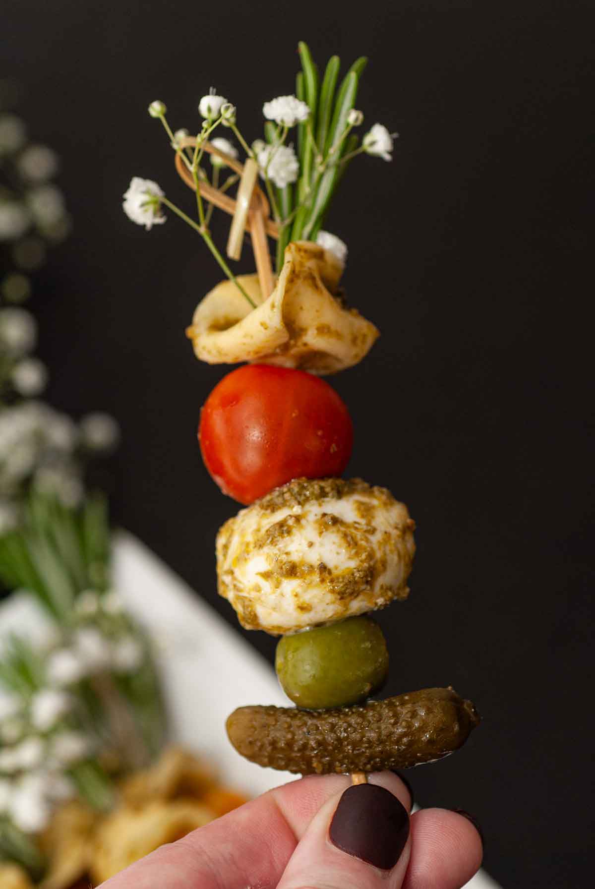 Fingers holding an antipasti skewer garnished with flowers and rosemary.