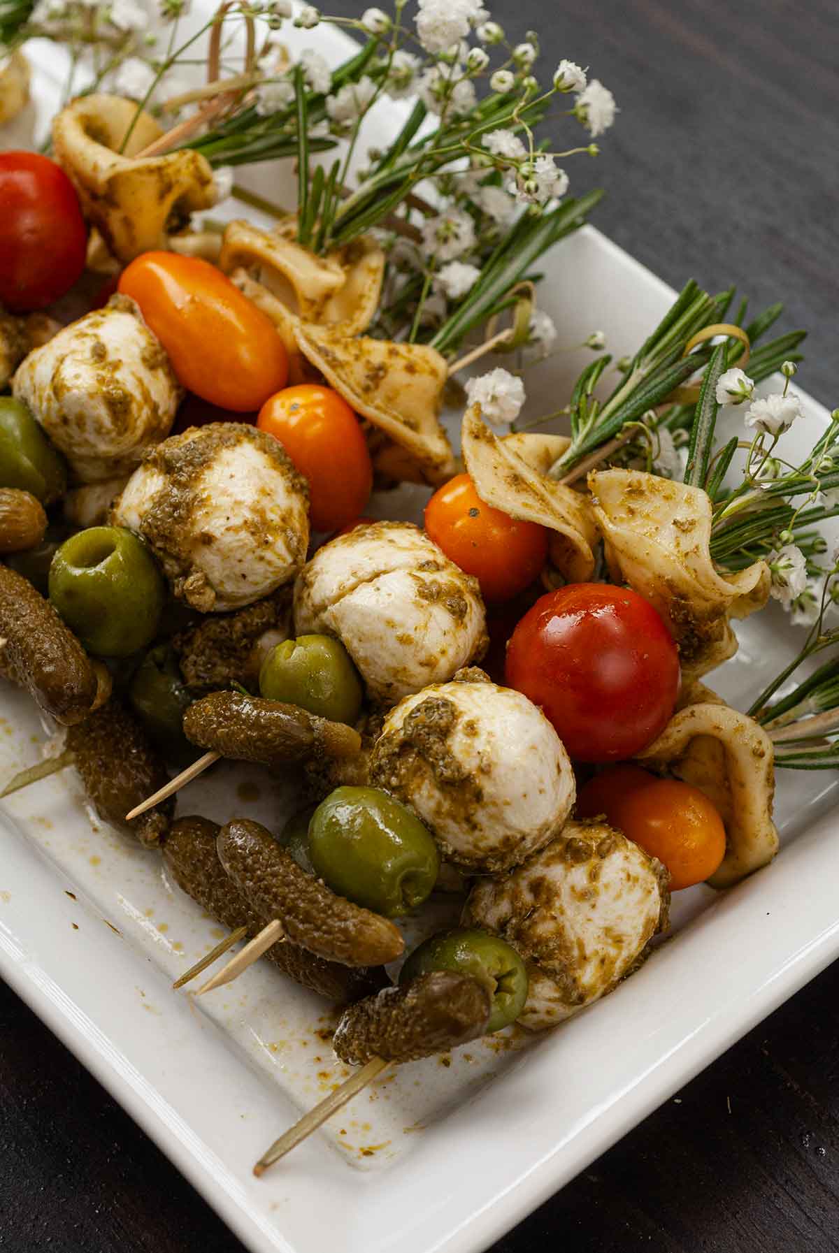 Antipasti skewers garnished with flowers and rosemary stacked on a plate on a wooden table.