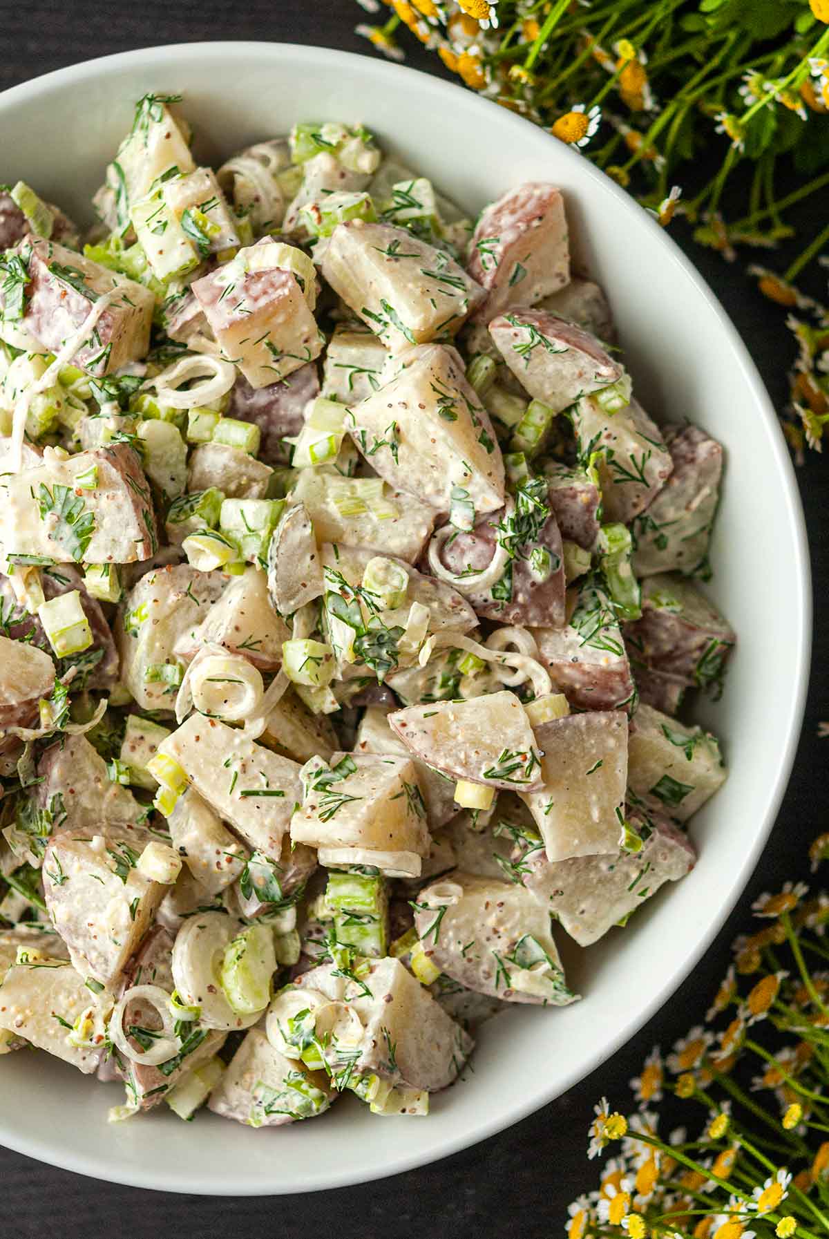 A bowl of dill potato salad on a table beside flowers.