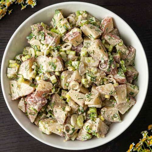 A bowl of dill potato salad centered on a table beside flowers.