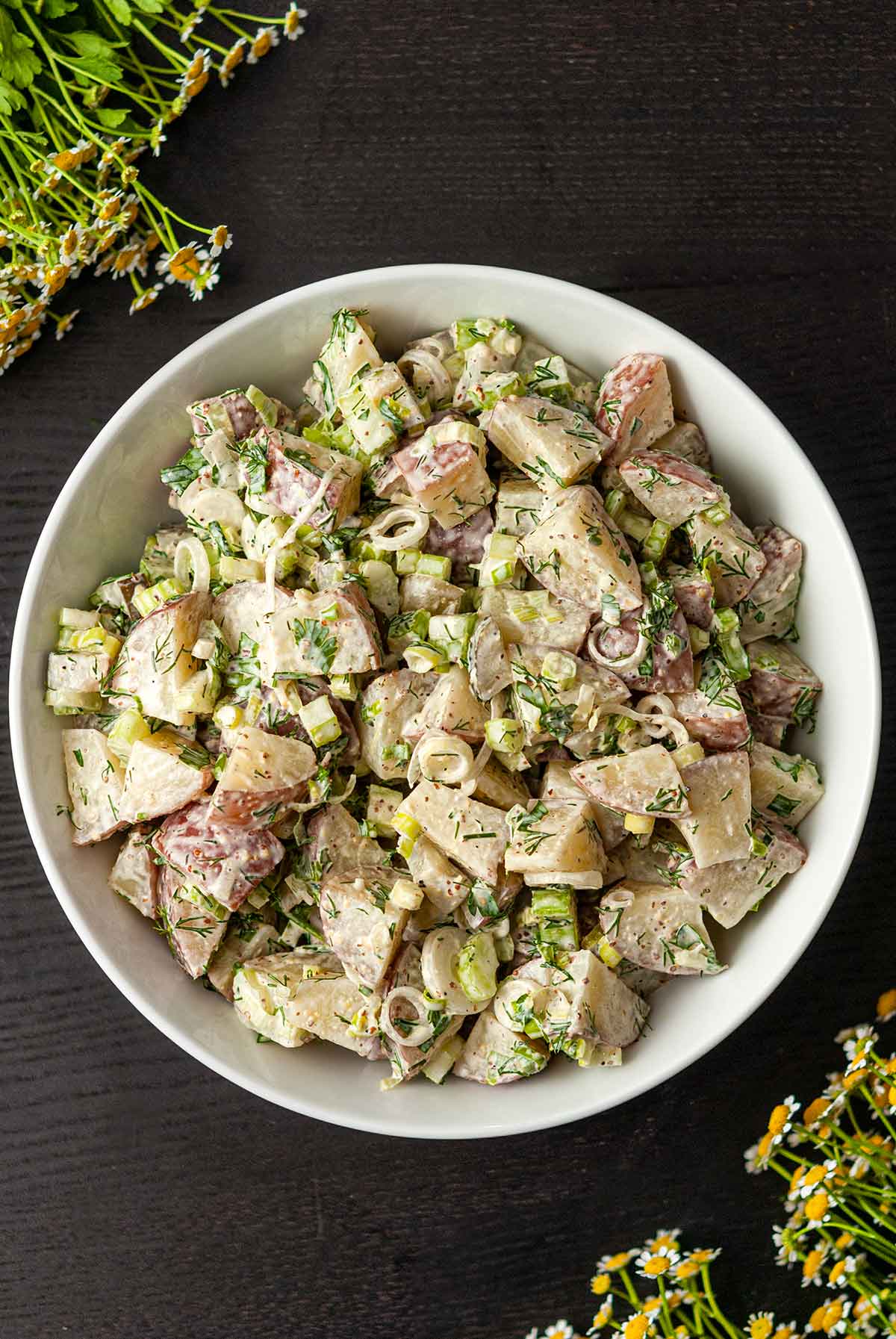 A bowl of dill potato salad centered on a table beside flowers.