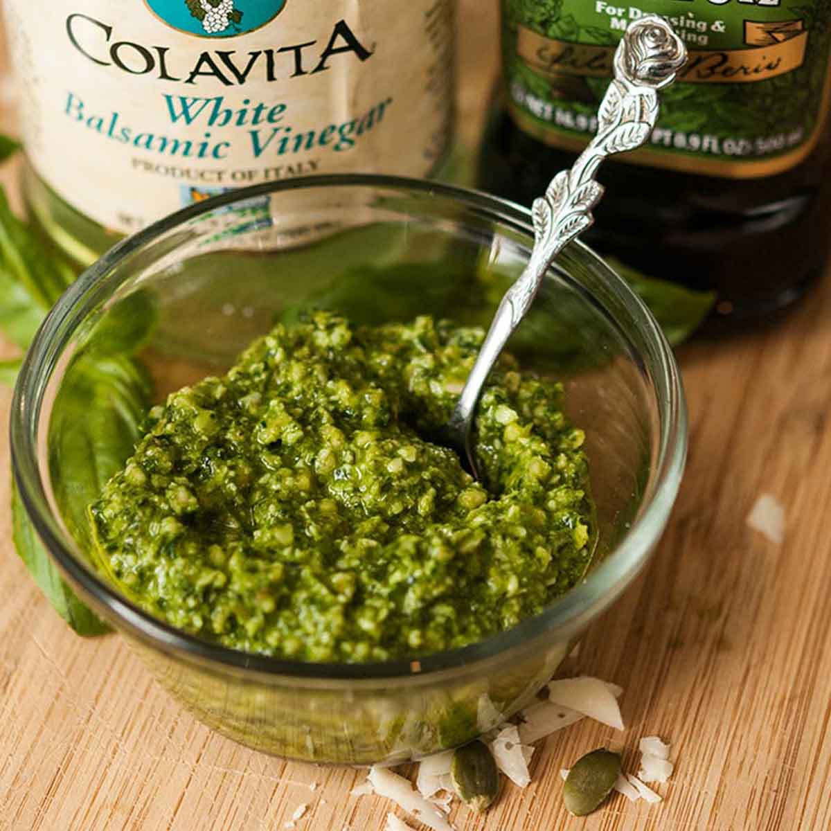 Homemade pesto in a bowl on a cutting board beside olive oil and balsamic vinigar.