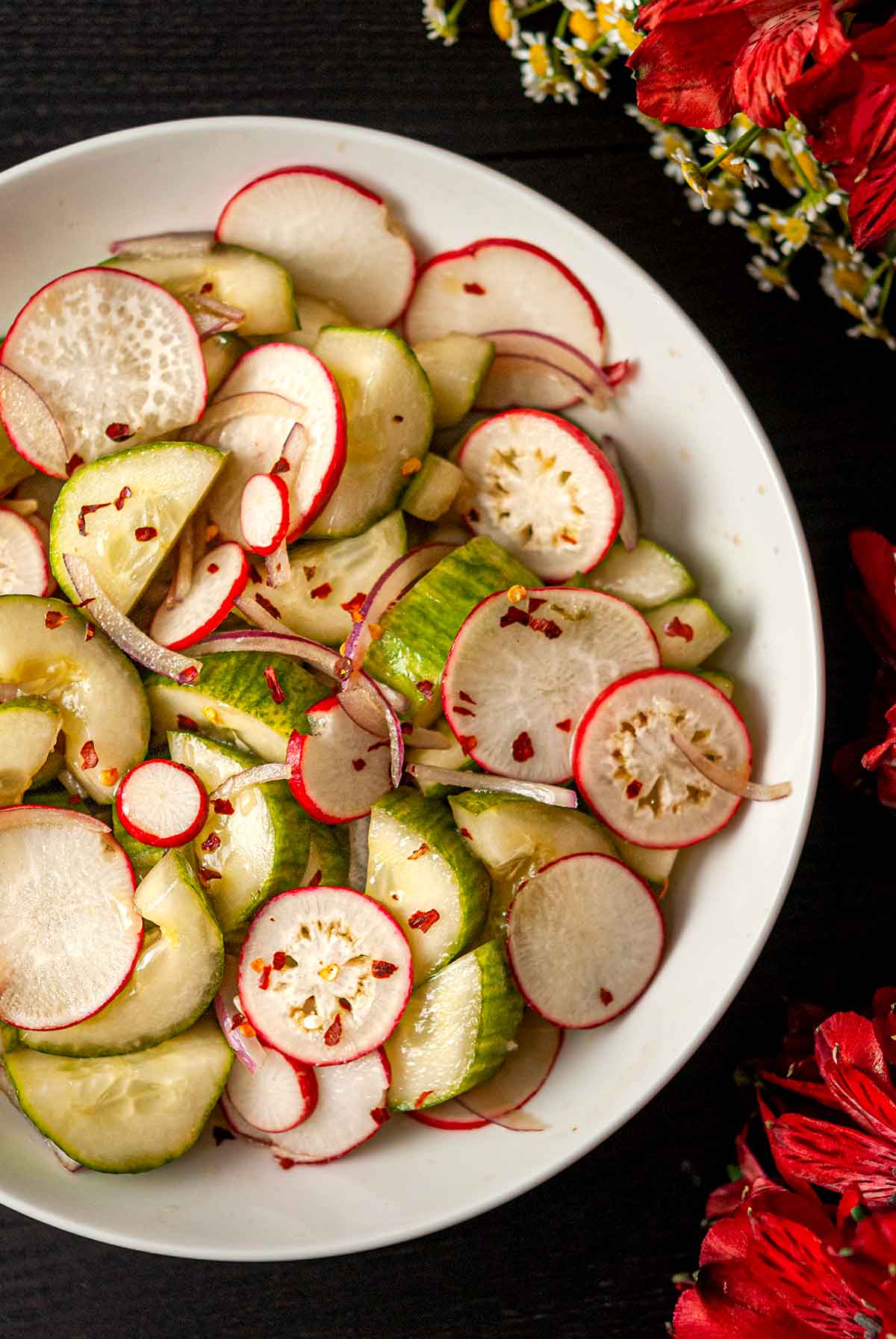 A bowl of ponzu cucumbers and radishes in a bowl beside flowers.