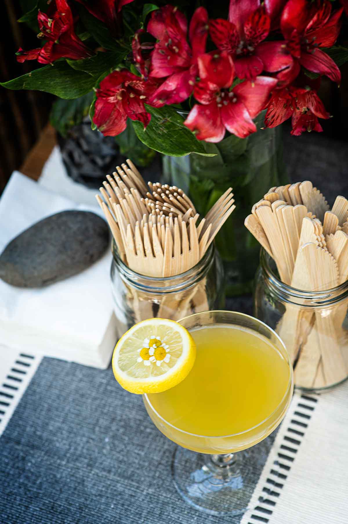 A lemon garnished cocktail on a table beside wooden knives, forks and flowers.