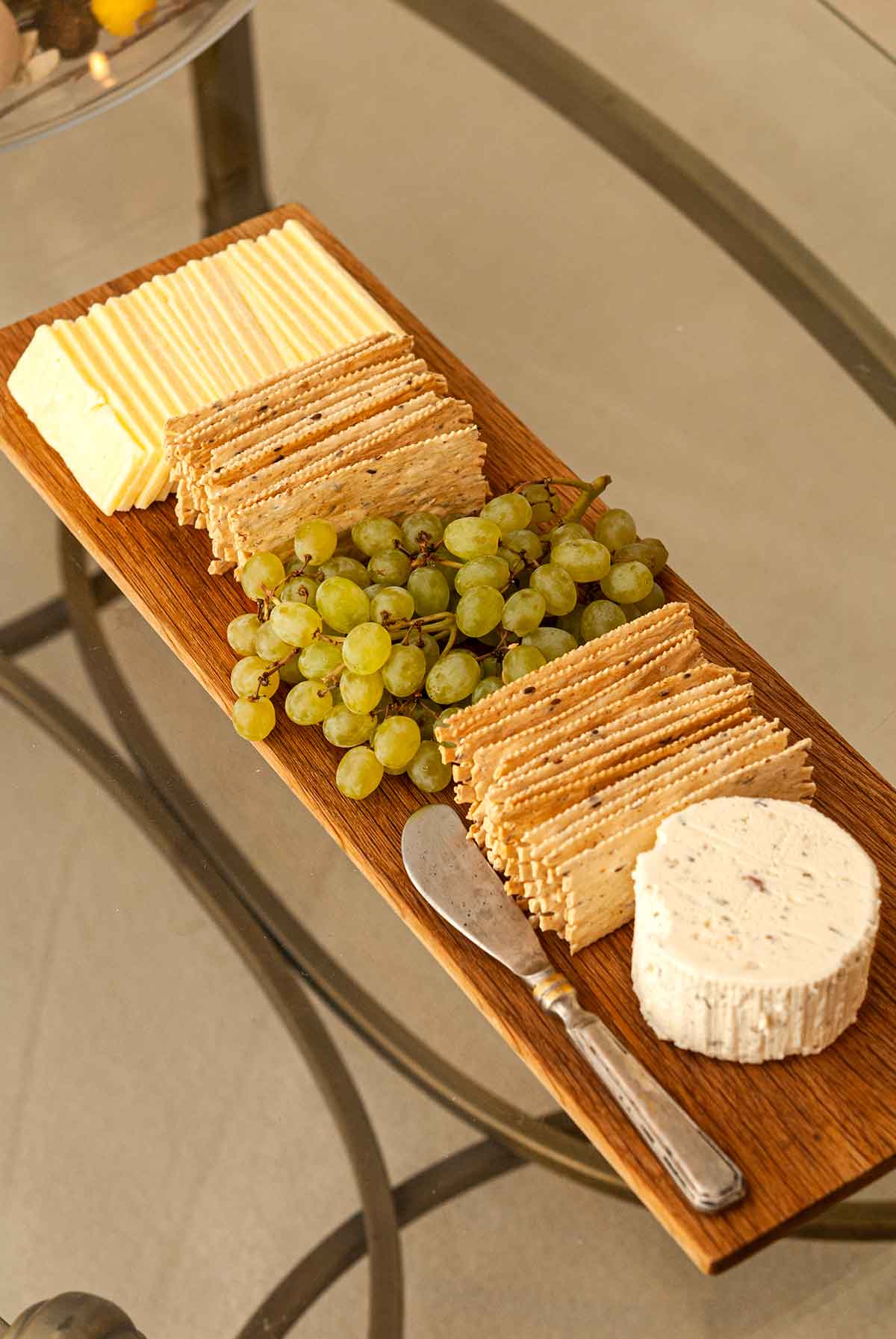 A board with 2 cheeses, crackers and small grapes on a glass table.