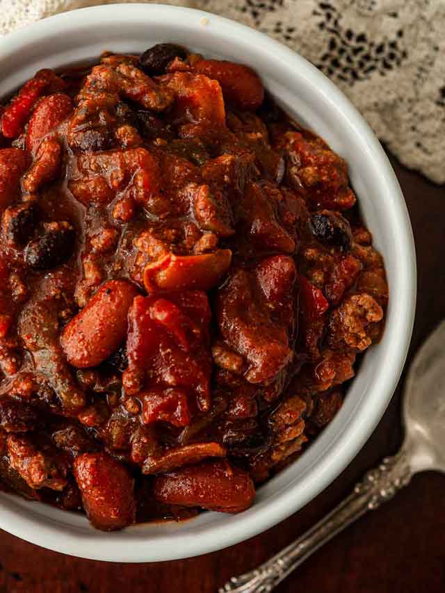A bowl of chili beside a spoon and a lace tablecloth.