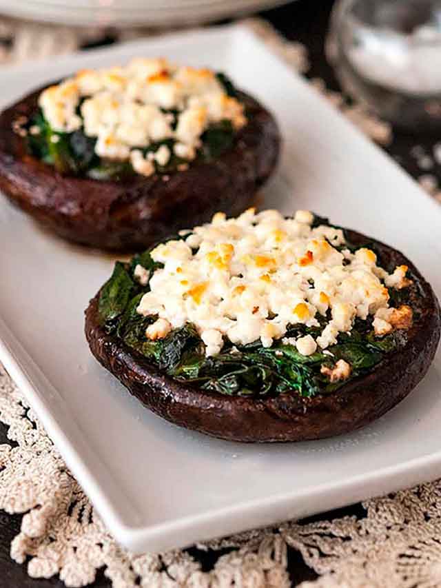 2 spinach and feta-stuffed mushroom caps on a white plate on a table, lined with lace with 2 plates in the background.