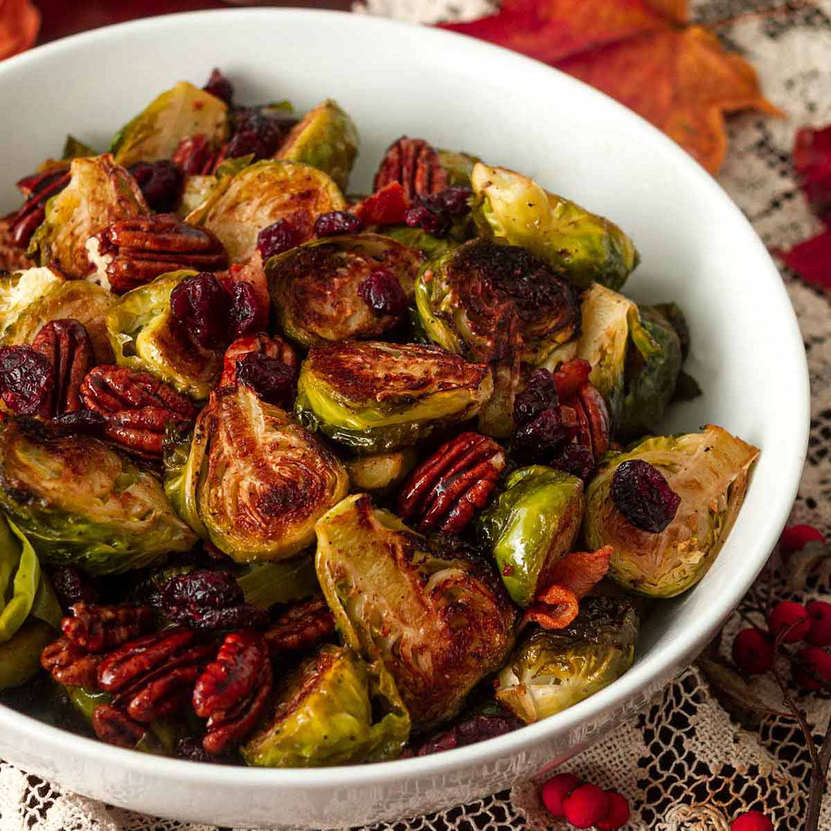 A bowl of Brussels sprouts, bacon, cranberries and pecans on a table with a lace table cloth and scattered leaves.