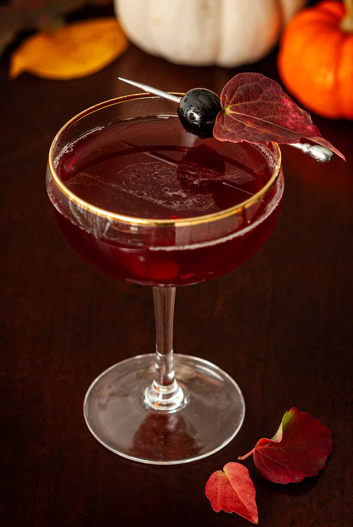 A red cocktail on a wooden table, garnished with a cherry and leaf, in front of pumpkins.