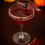 A red cocktail on a wooden table, garnished with a cherry and leaf, in front of pumpkins.