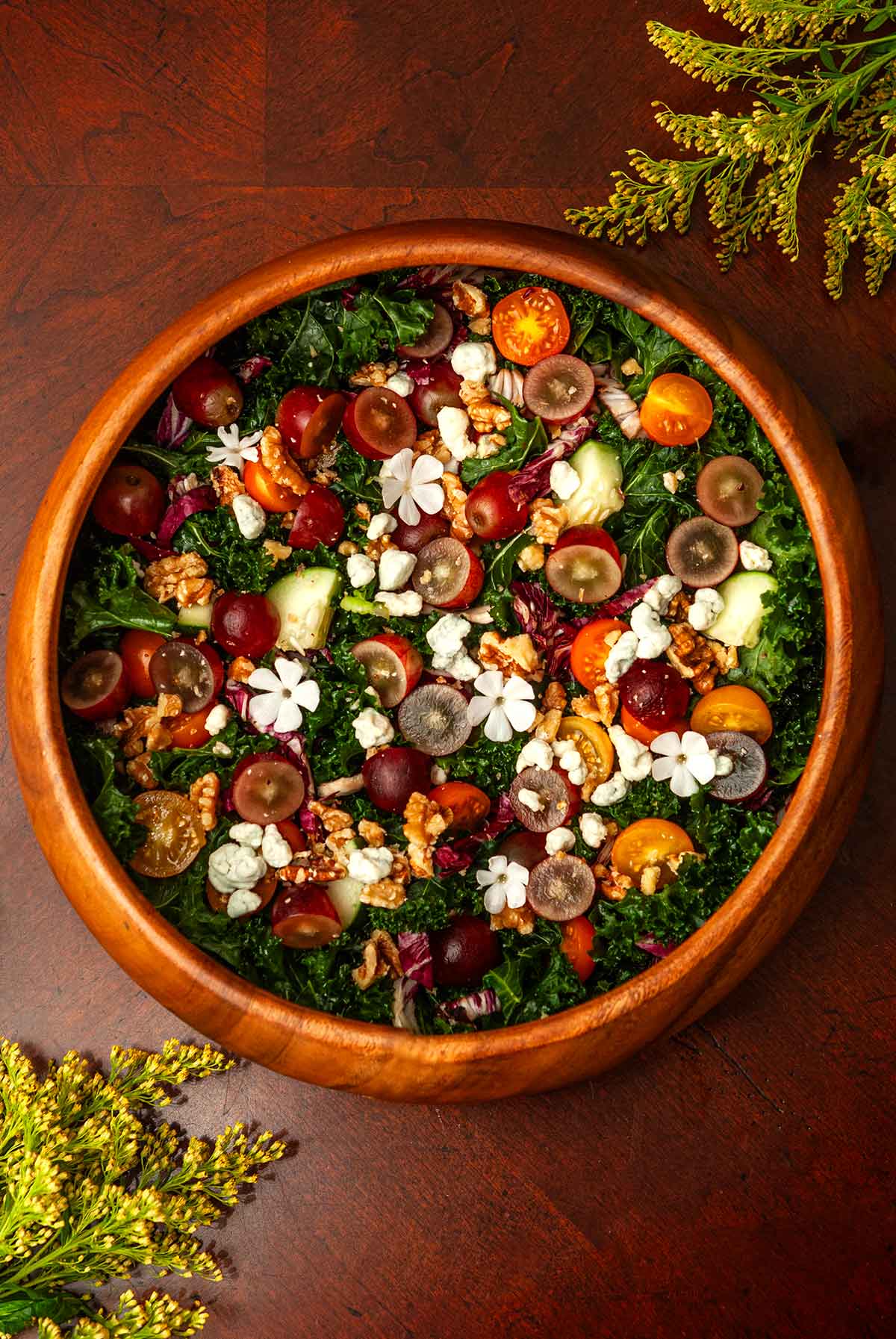 A wooden bowl of salad with grapes on a table with flowers.