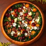 A wooden bowl of salad with grapes on a table with flowers.