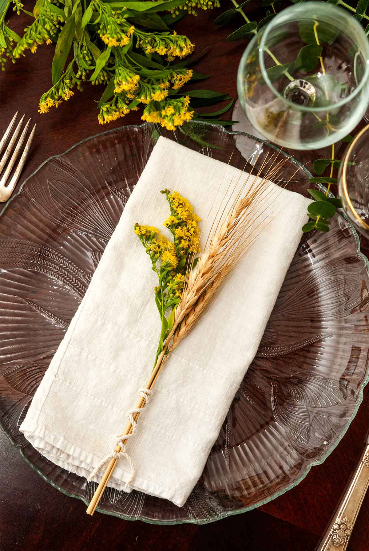 A small bundle of goldenrod and wheat on a napkin on a plate on a table with glasses.