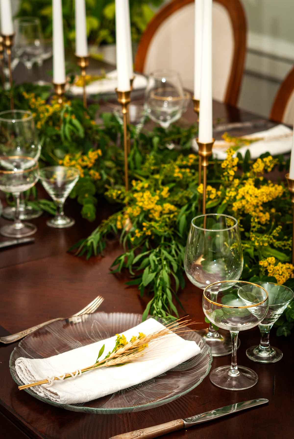 A eucalyptus and goldenrod centerpiece with candle sticks, plates and glasses on a table.