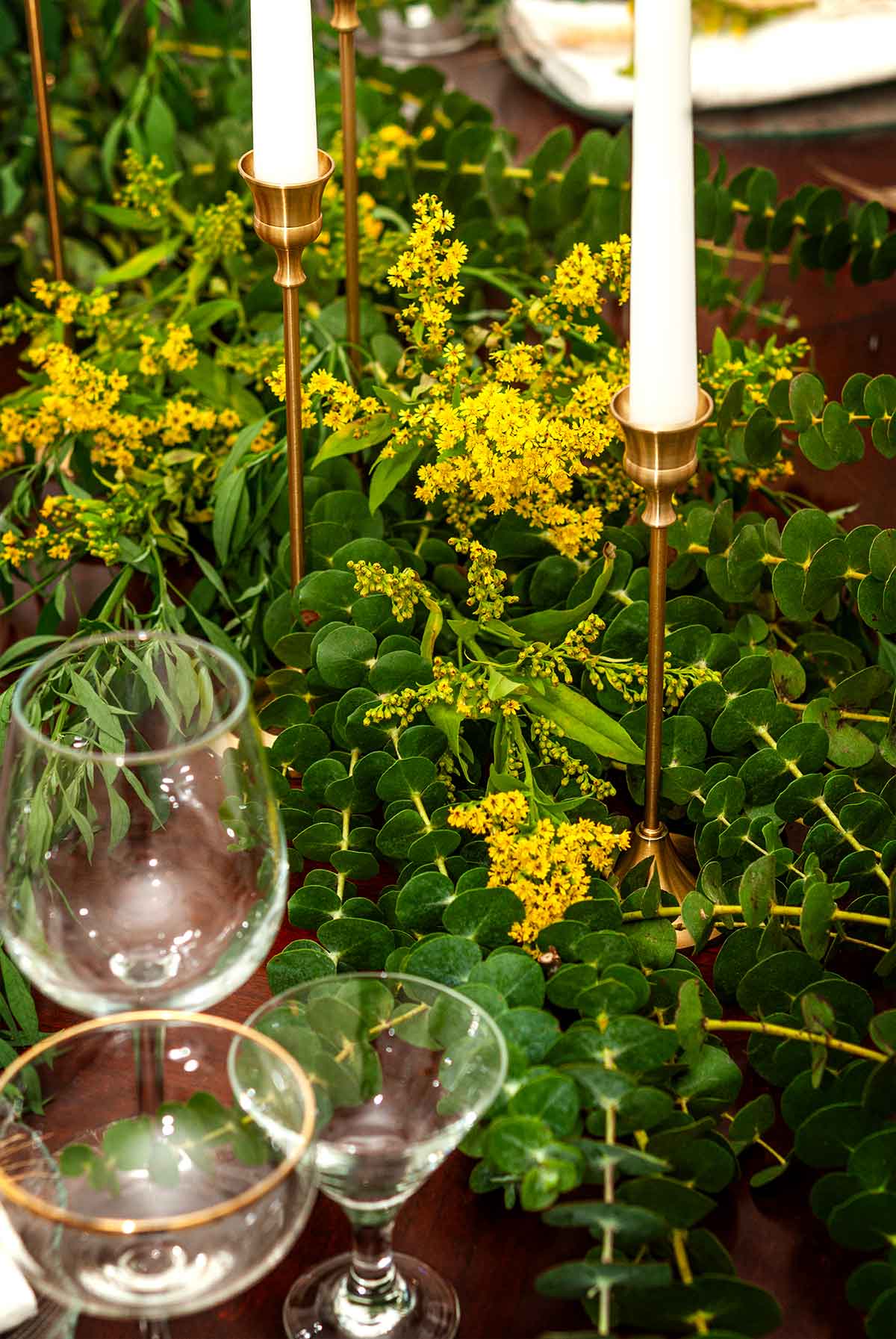 A eucalyptus and goldenrod centerpiece with candle sticks and glasses on a table.