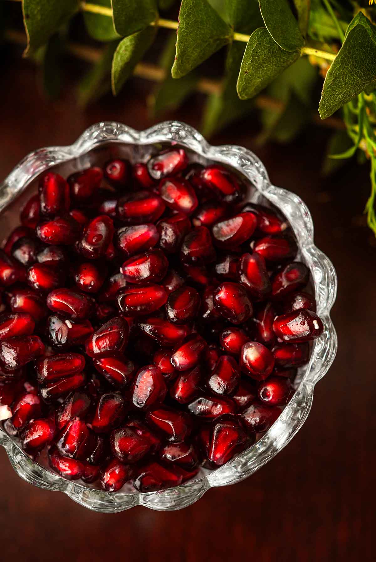 A bowl of pomegranate seeds on a table beside sprigs of eucalyptus. 