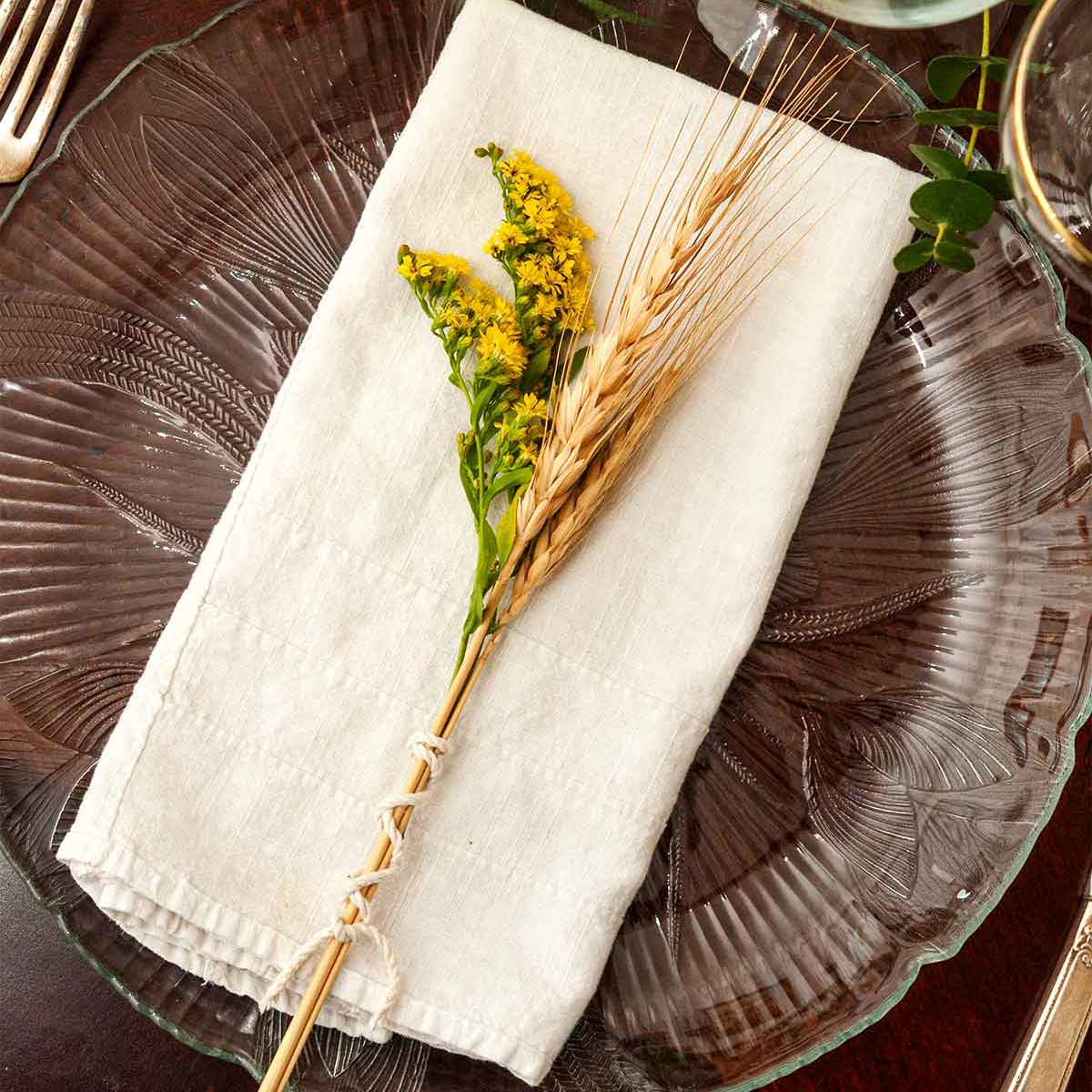 A small bundle of goldenrod and wheat on a napkin on a plate on a table with glasses.