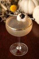 A cocktail with a lychee eyeball garnish beside 2 white pumpkins and flowers on a table.