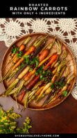 A wooden platter of roasted carrots, garnished with parsley and pomegranate seeds on a lace tablecloth.