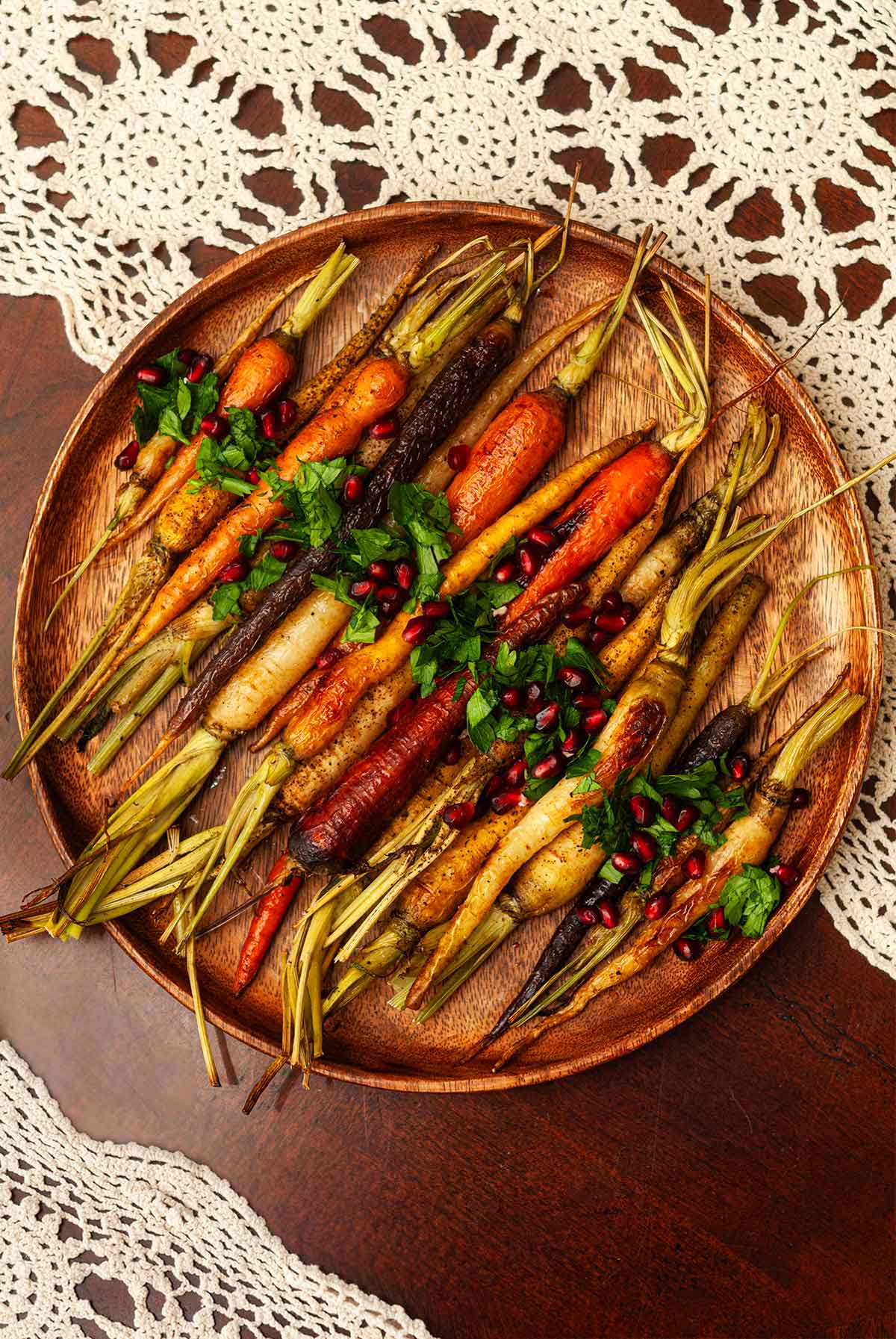 A wooden platter of roasted carrots, garnished with parsley and pomegranate seeds on a lace tablecloth.