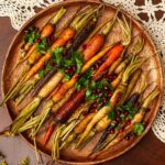 A wooden platter of roasted carrots, garnished with parsley and pomegranate seeds on a lace tablecloth.