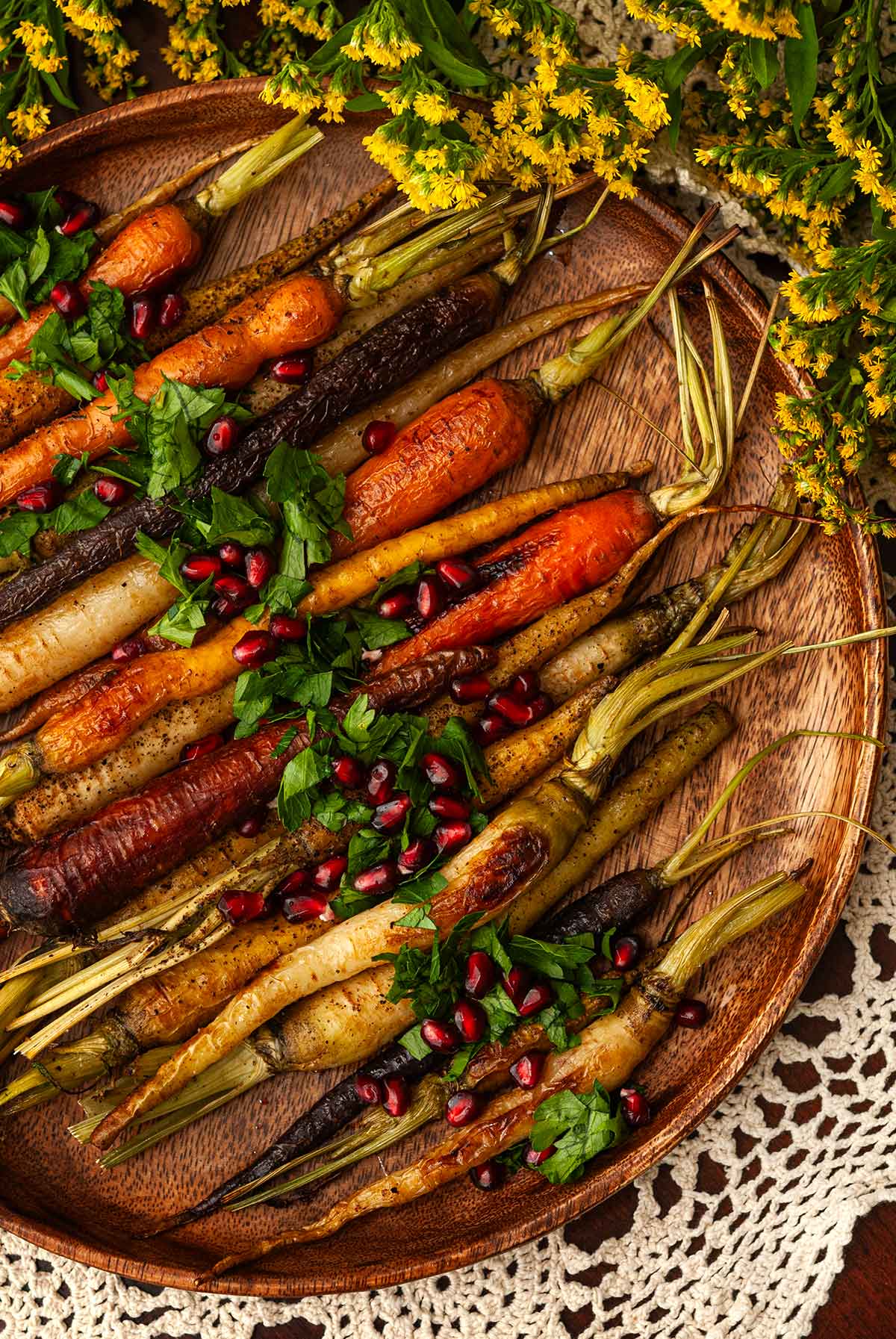 A wooden platter of roasted carrots on a lace tablecloth beside flowers..