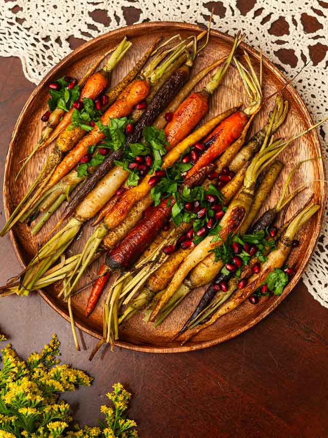 A wooden platter of roasted carrots, garnished with parsley and pomegranate seeds on a lace tablecloth.