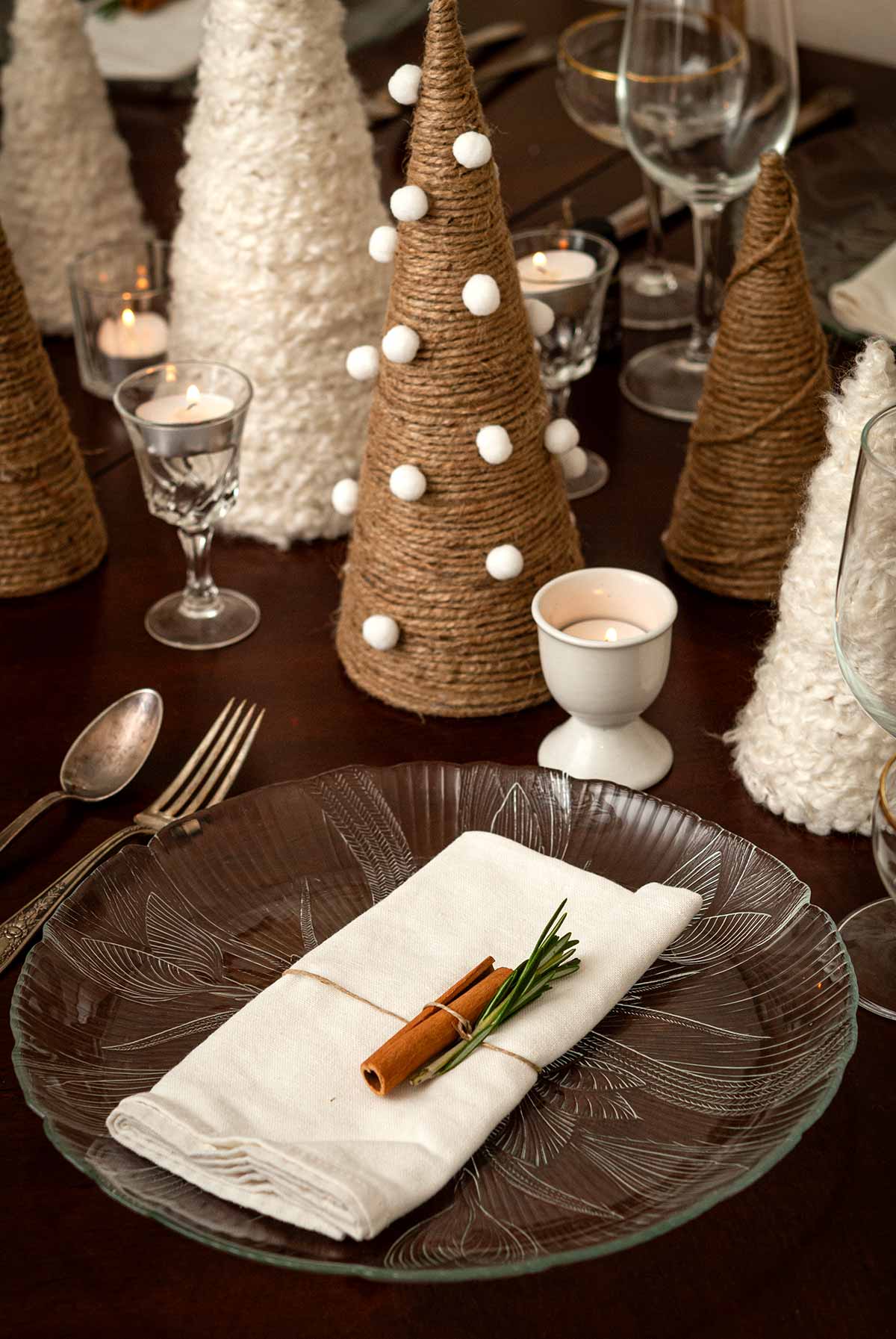 A place setting with a cinnamon stick and rosemary in front of wool and twine trees on a table.