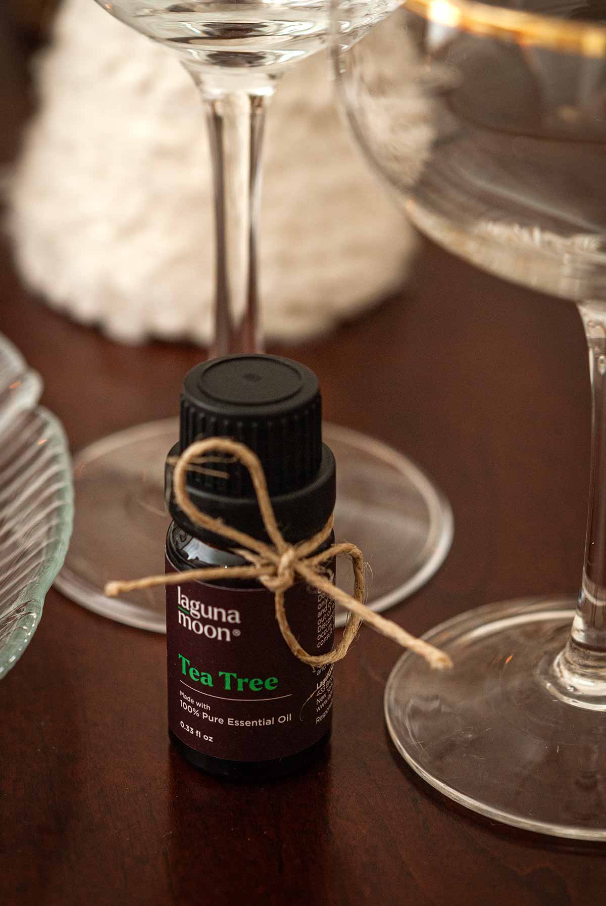 A small bottle of tee tree oil tied with a little string on a table i front of glassware.