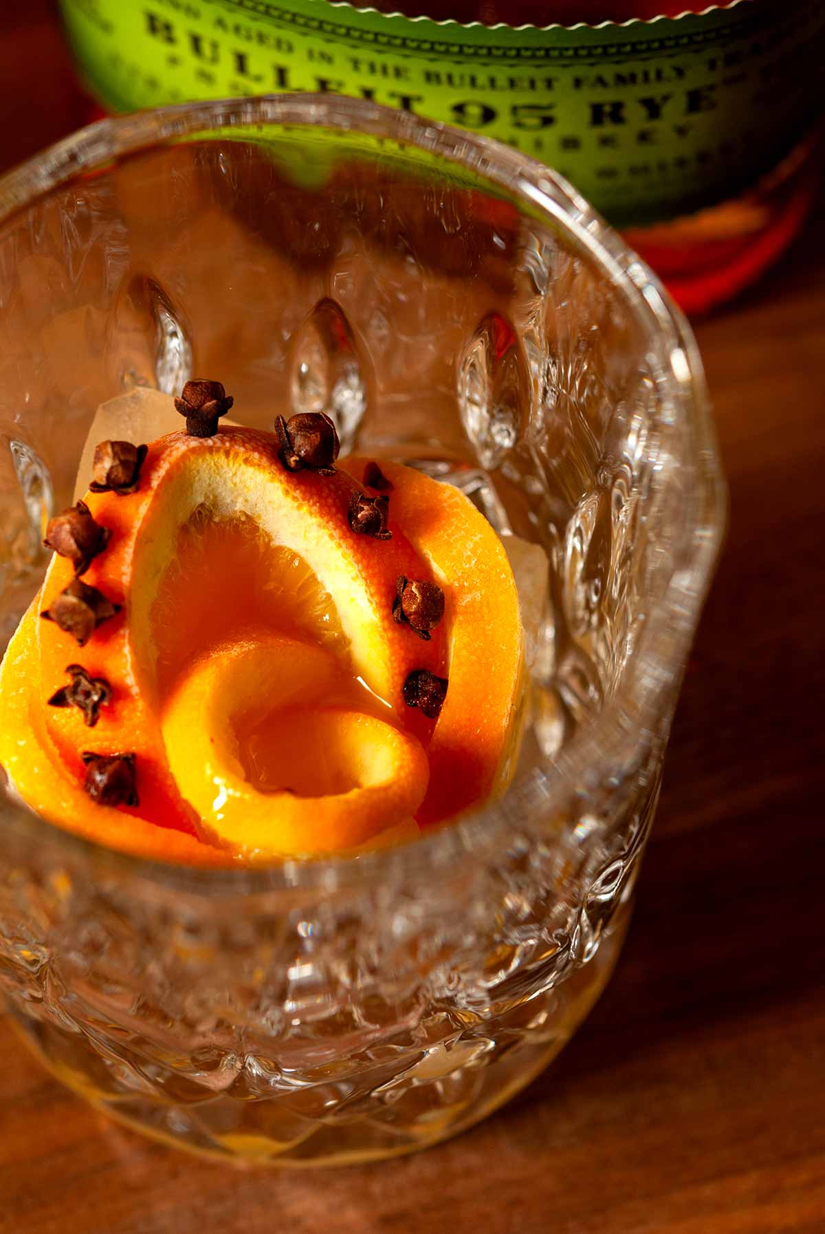 A cocktail glass with a orange-clove rose ice cube on a table beside a bottle of whisky.