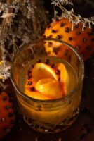 A cocktail in front of a clove orange with a clove orange rose ice cube surrounded by flowers.