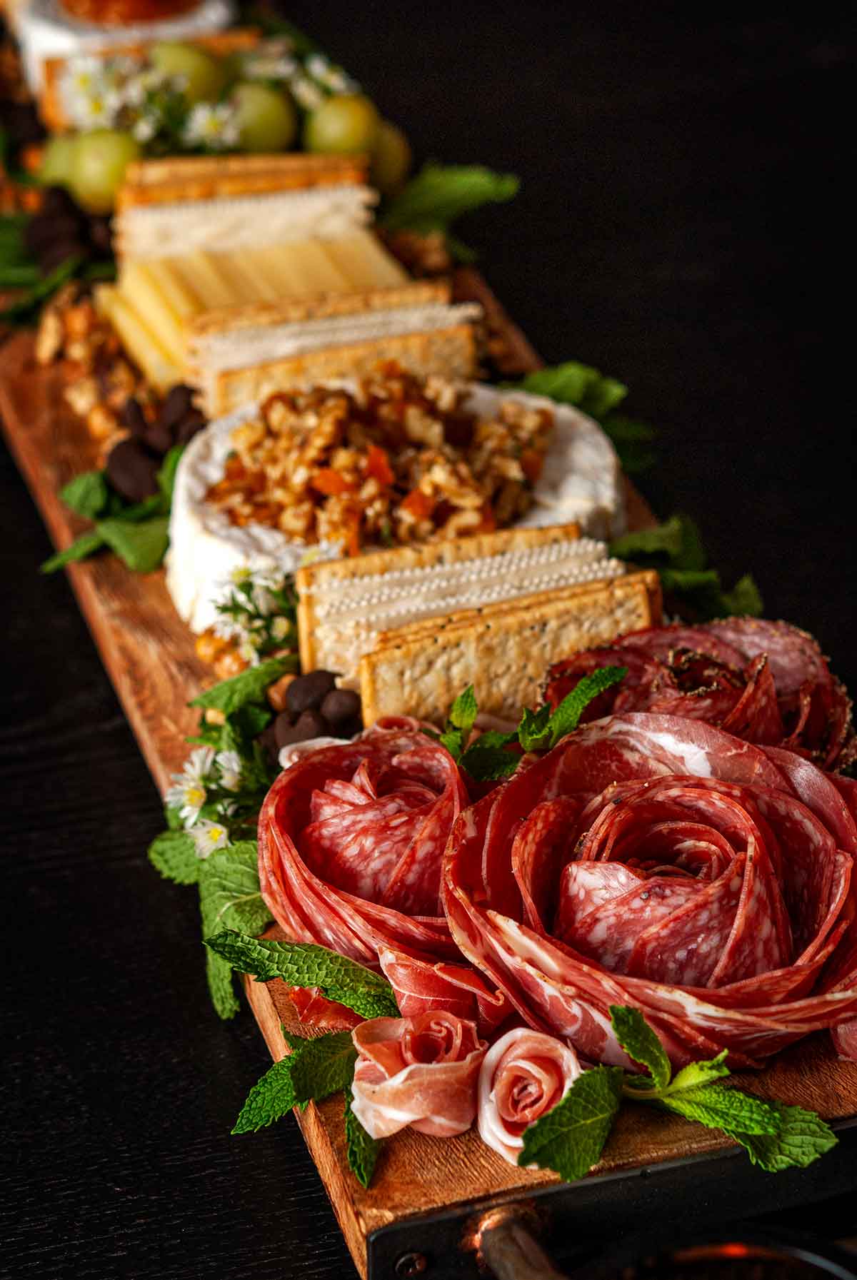 An ornate cheese board with meat roses, 2 cheeses, crackers and grapes.