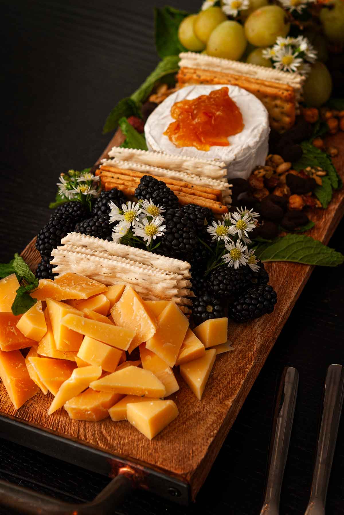An ornate cheeseboard with 2 cheeses, berries, grapes, crackers and flowers.