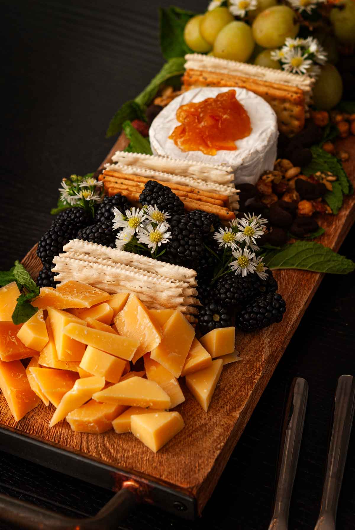 An ornate cheeseboard with 2 cheeses, berries, grapes, crackers and flowers.