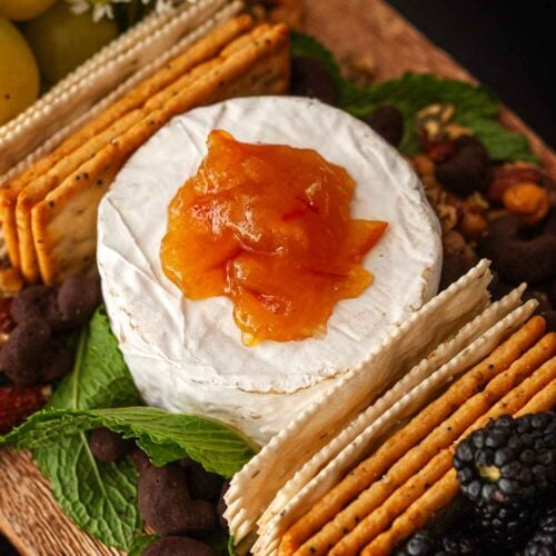 Cheese on a board, topped with marmalade, surrounded by crackrs and mint.