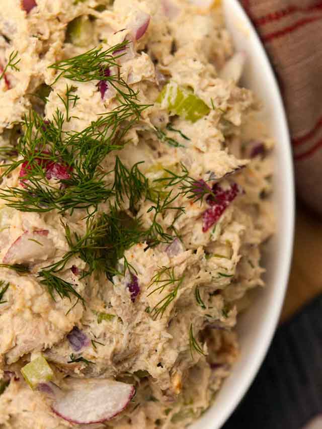 A bowl of chicken salad, topped with dill beside a vintage tea towel.