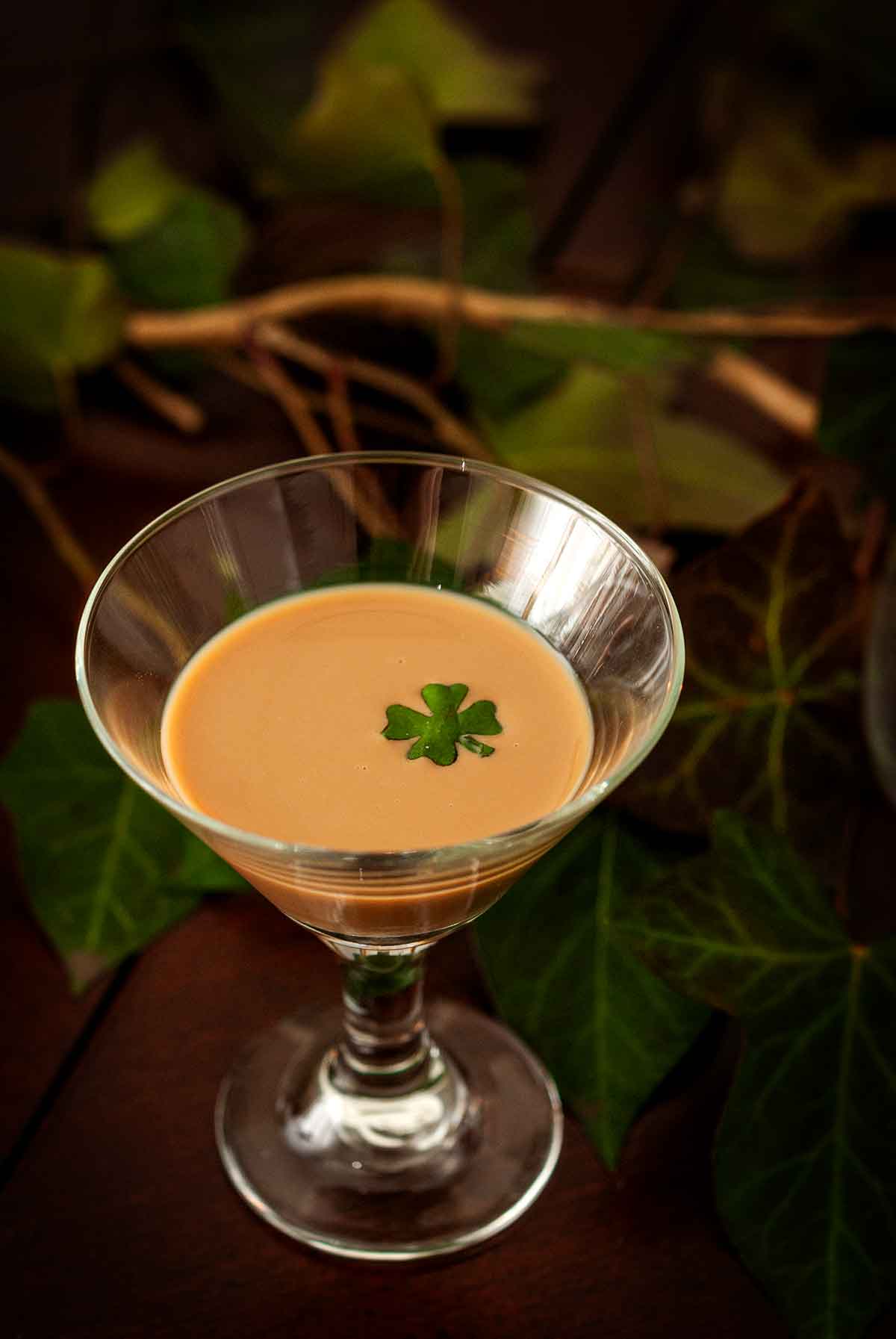 A small glass of a cream cocktail garnished with a basil clover, surrounded by ivy.