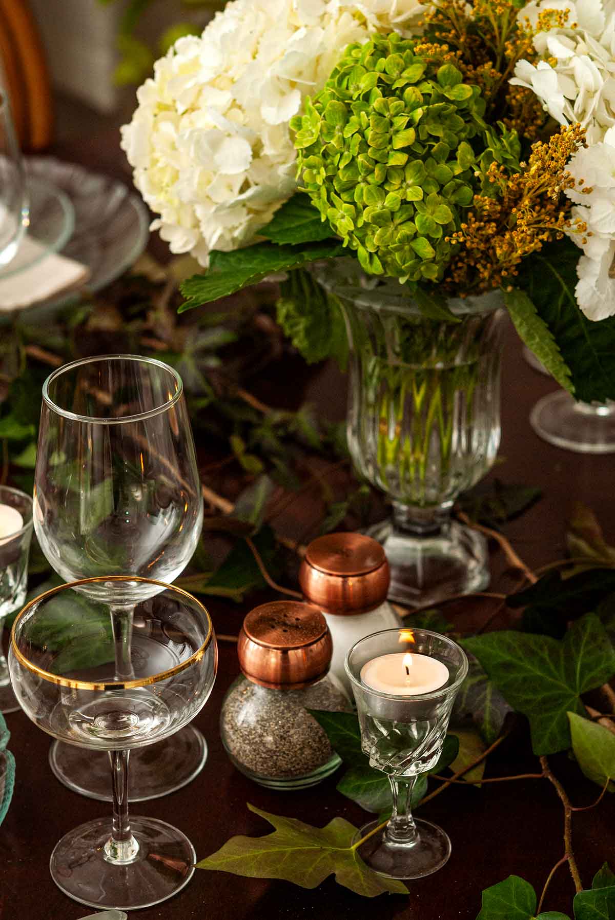 A vase of flowers, salt and pepper shakers, glassware and a candle in a cordial glass on a table.