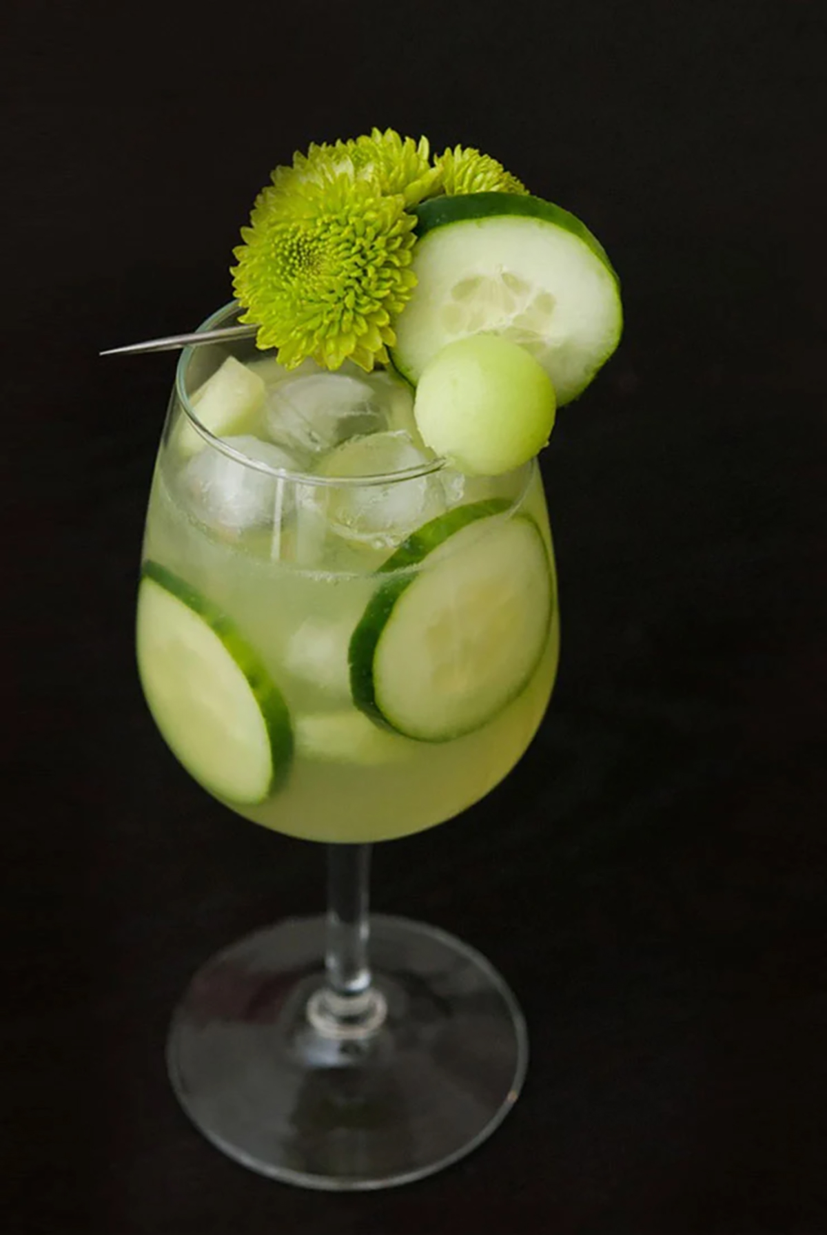 A green cocktail on a black table, garnished with aren flowers, cucumber and a ball of honeydew with cucumbers in the glass.