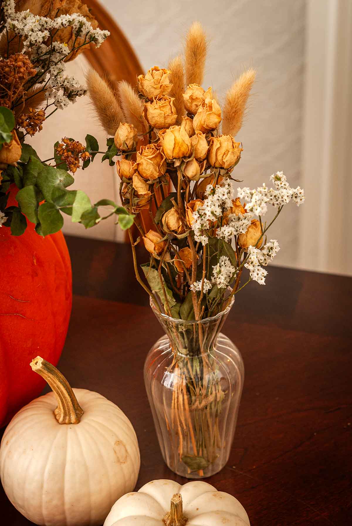 A small glass vase with dry flowers beside 3 pumpkins.