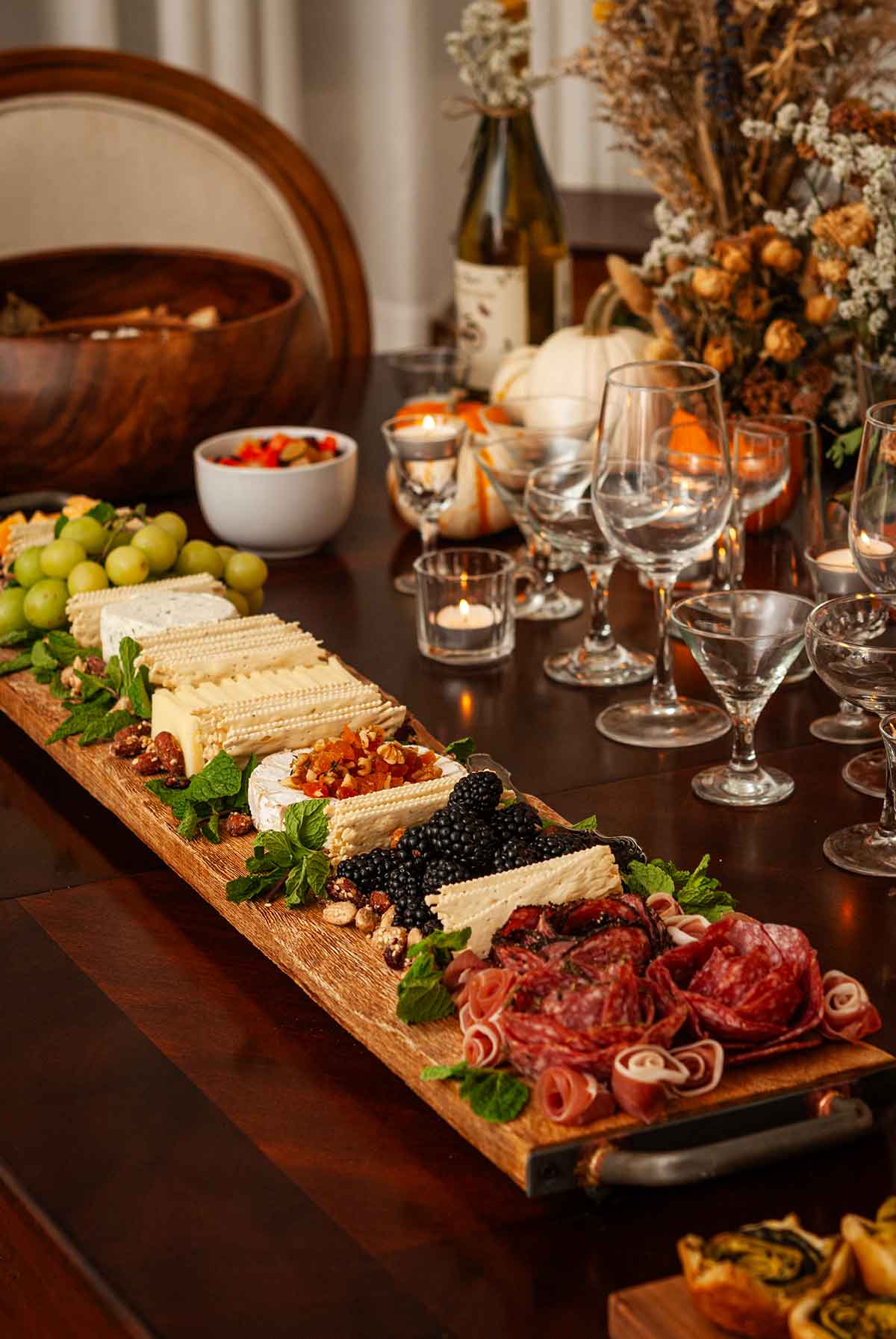A long cheeseboard with assorted snacks on a table with glasses and pumpkin bouquets.